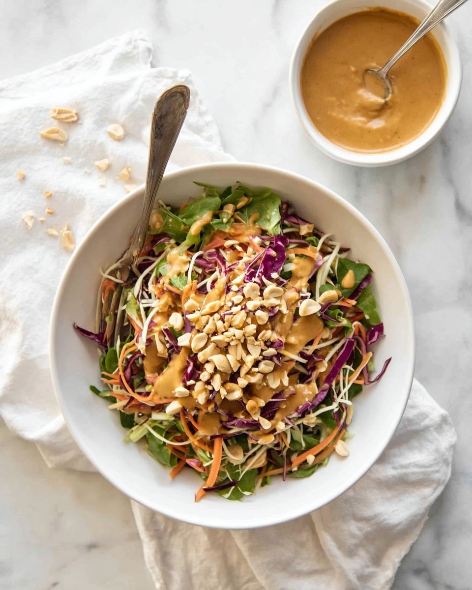 A bowl filled with a colorful salad that has several layers: at the bottom are thin green strips of leafy vegetables, followed by shredded purple cabbage and bright orange carrot strips, topped with light yellow crunchy sticks and sliced green onions. On top, there is a drizzle of smooth, light brown peanut sauce and a small pile of chopped peanuts. A spoon is placed inside the bowl, which sits on a white marbled surface with a white cloth nearby. Next to the bowl is a white bowl of extra peanut sauce with chopped peanuts and a spoon inside. photo taken with an iphone --ar 4:5 --v 7