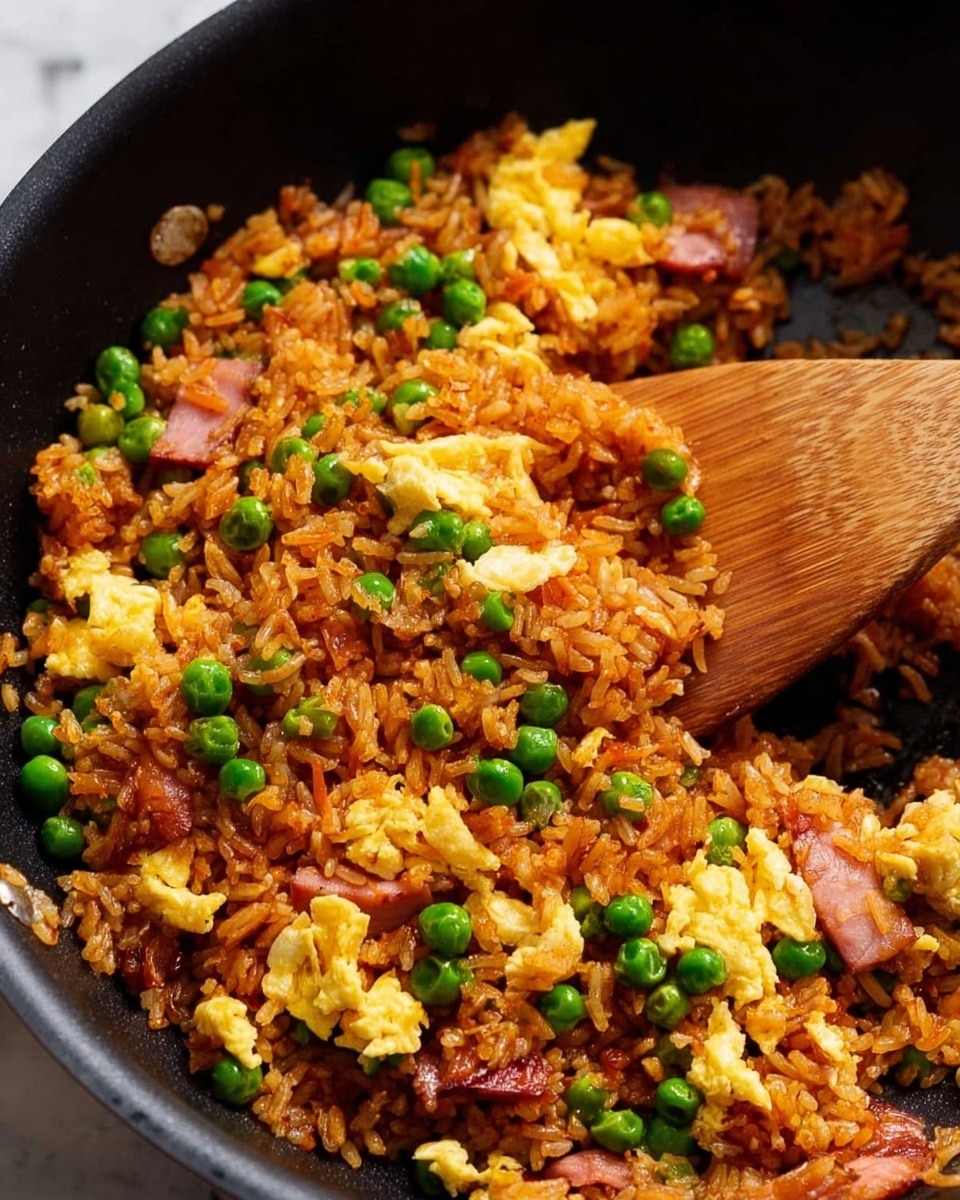 The image shows a close-up of fried rice in a black pan, with one layer of reddish-brown cooked rice mixed with peas, scrambled eggs, and small pieces of meat, spread evenly throughout. The peas are bright green, scattered all over the rice, while the scrambled eggs are yellow and fluffy, broken into chunks. The meat pieces are small and dark pink. A wooden spatula is partially visible on the right side, scooping some rice. The background is a white marbled texture. photo taken with an iphone --ar 4:5 --v 7