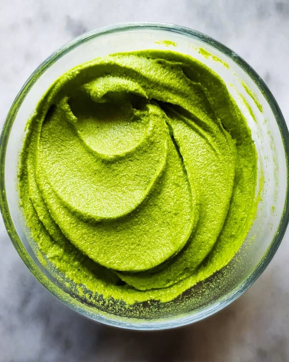 A close-up view of a clear glass bowl filled with smooth bright green paste, swirled neatly in a circular pattern with a creamy texture and no visible chunks, placed on a white marbled surface photo taken with an iphone --ar 4:5 --v 7