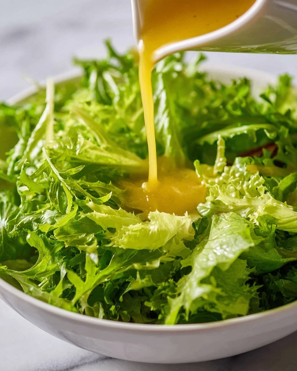 A close-up shot shows bright green curly lettuce leaves filling a white bowl, with a light yellow dressing being poured over the salad from a small container just above. The lettuce leaves are fresh and crisp, with some parts catching light, showing their texture and edges clearly. The background is a soft white marbled texture, blurred to keep focus on the salad. Photo taken with an iphone --ar 4:5 --v 7