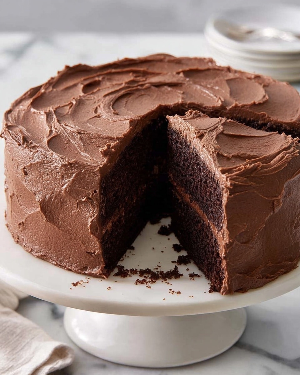 A round chocolate cake with two thick layers of dark brown, moist sponge separated by a middle layer of smooth chocolate frosting. The entire cake is covered in a thick, even layer of creamy chocolate frosting with soft swirls and subtle texture on the top and sides. One large slice is cut and slightly pulled out, revealing the inside layers clearly. The cake sits on a white pedestal cake stand, placed on a white marbled surface. Photo taken with an iphone --ar 4:5 --v 7