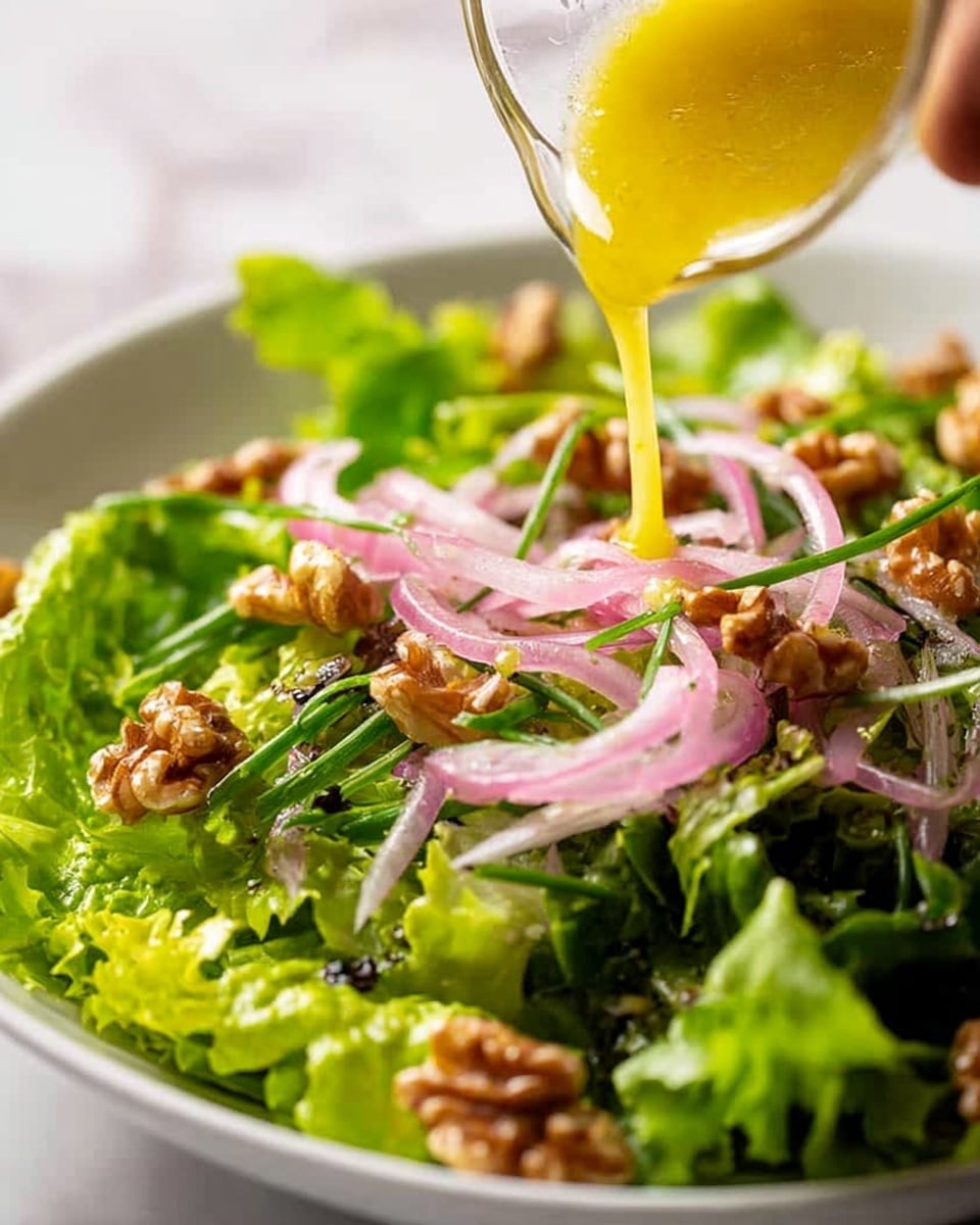 A close-up view of a fresh green salad in a shallow white bowl, showing several layers of leafy lettuce with deep green and light green colors. The salad is sprinkled with thin, curled pale pink onion slices, long dark green chives, and pieces of light brown walnuts scattered on top. A woman's hand is pouring a yellow dressing in a thin stream over the salad, making some leaves glisten. The background shows a white marbled texture. photo taken with an iphone --ar 4:5 --v 7