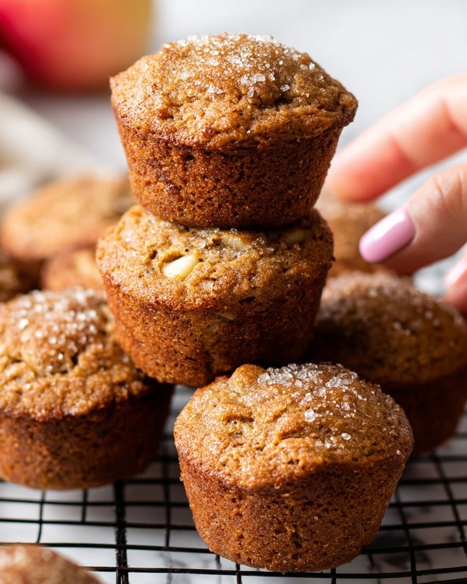 The image shows a close-up of five golden-brown muffins with a slightly rough texture, sitting stacked on a black cooling rack. Each muffin has small pieces of light yellow fruit or nuts visible inside, and a light sprinkle of coarse sugar crystals on top, adding a bit of sparkle. The muffins have rounded, slightly domed tops and moist interiors visible through the crumbly crust. The background surface is a white marbled texture, and there is a woman's hand visible in the lower left corner holding a slice of fruit. The overall mood is warm and fresh. photo taken with an iphone --ar 4:5 --v 7