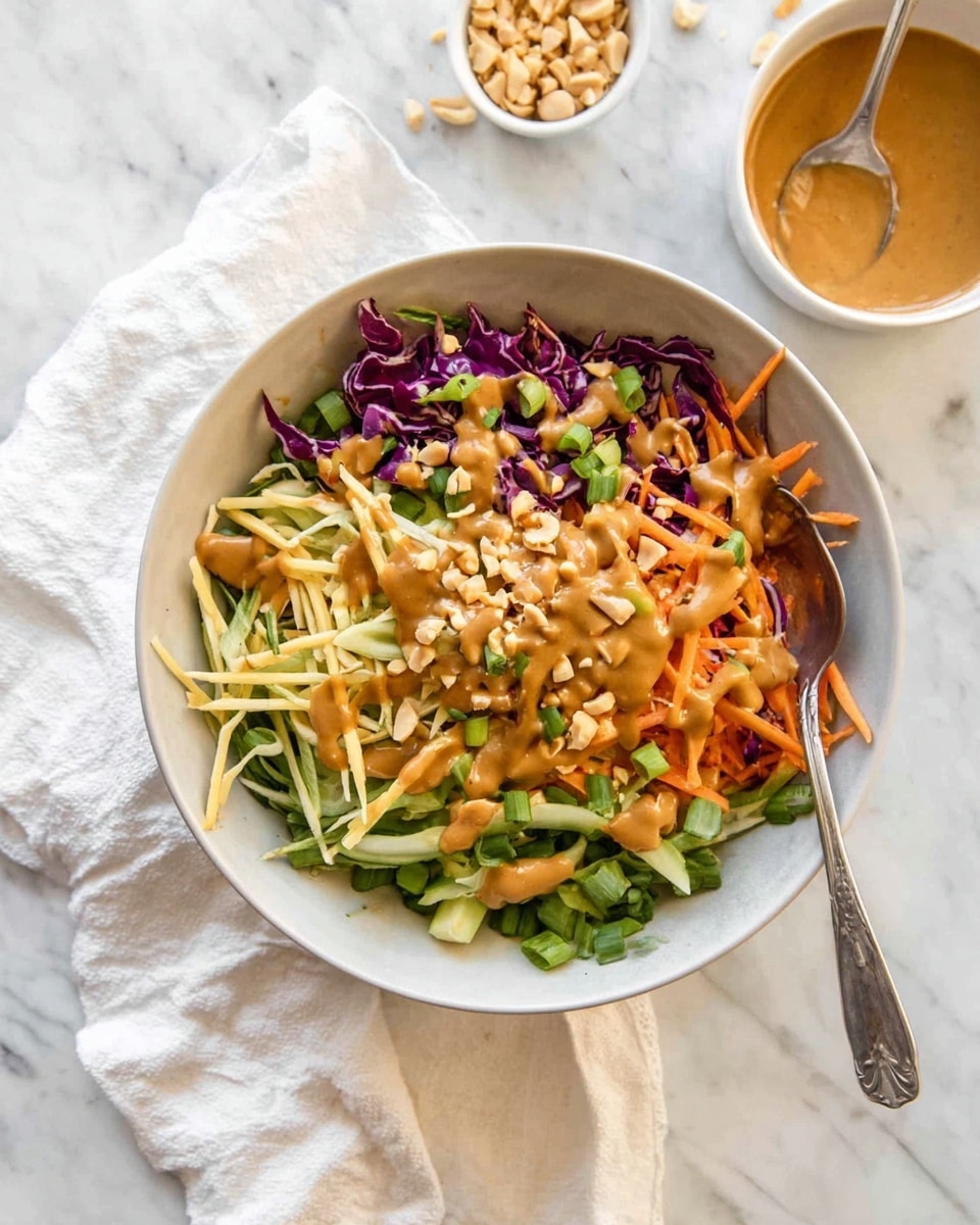A white bowl filled with a colorful salad featuring a mix of thin shredded layers: green leafy strips, purple cabbage, thin orange carrot sticks, and light-colored crunchy noodles. The salad is topped with a drizzle of creamy tan dressing and chopped peanuts in the center. A silver fork is placed inside the bowl, resting against the side. To the top right, a small white bowl contains more of the tan dressing with peanut pieces inside, with a silver spoon resting in it. The entire setting is on a white marbled surface with a white cloth partially under the bowl. Photo taken with an iphone --ar 4:5 --v 7