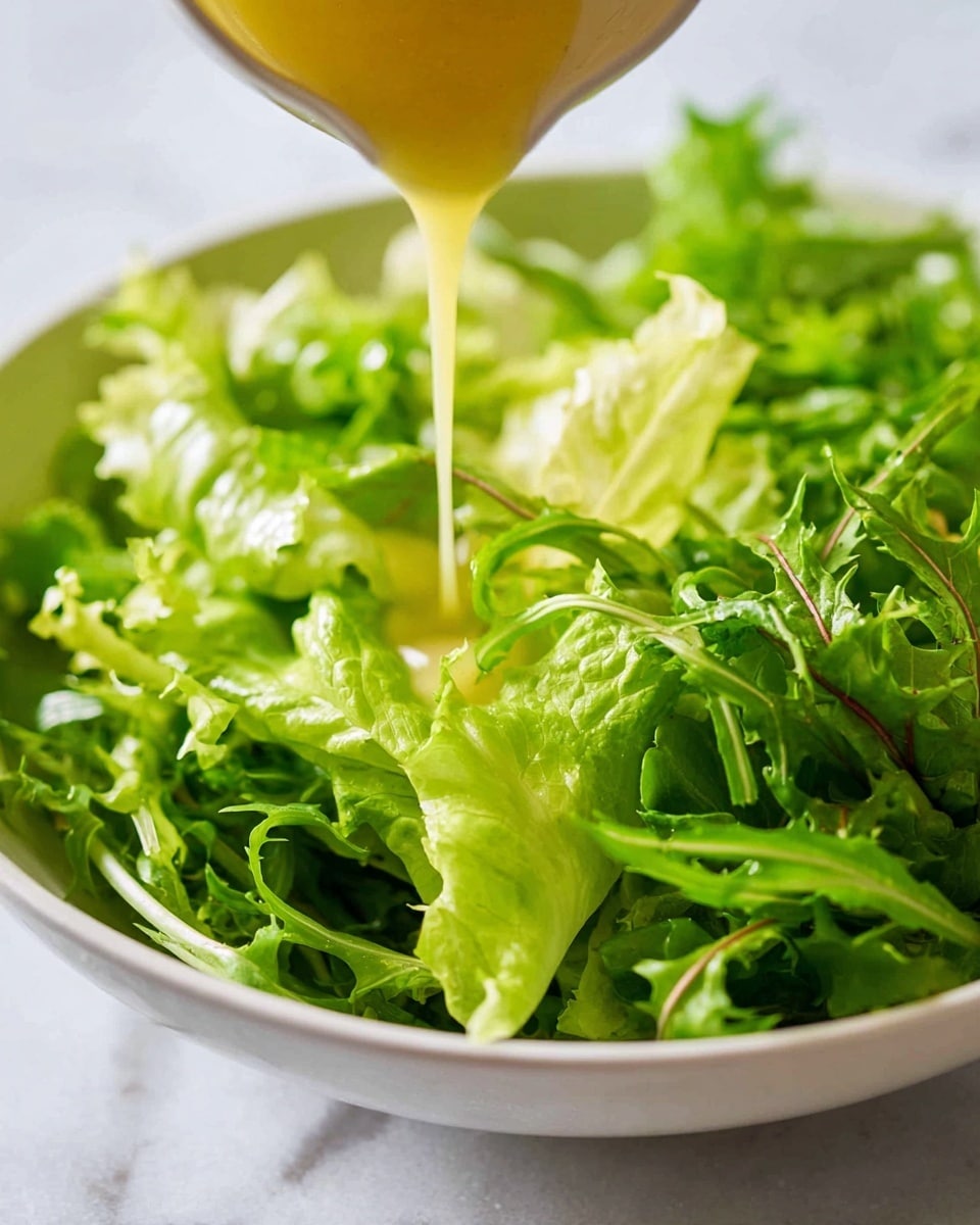 A close-up view of fresh, bright green leafy lettuce piled in a white bowl, with a thick, smooth golden-yellow dressing being poured from a small white pitcher onto the center of the leaves, highlighting the texture of the dressing as it drips and flows over the curly edges of the lettuce. The background shows a soft, white marbled texture. photo taken with an iphone --ar 4:5 --v 7