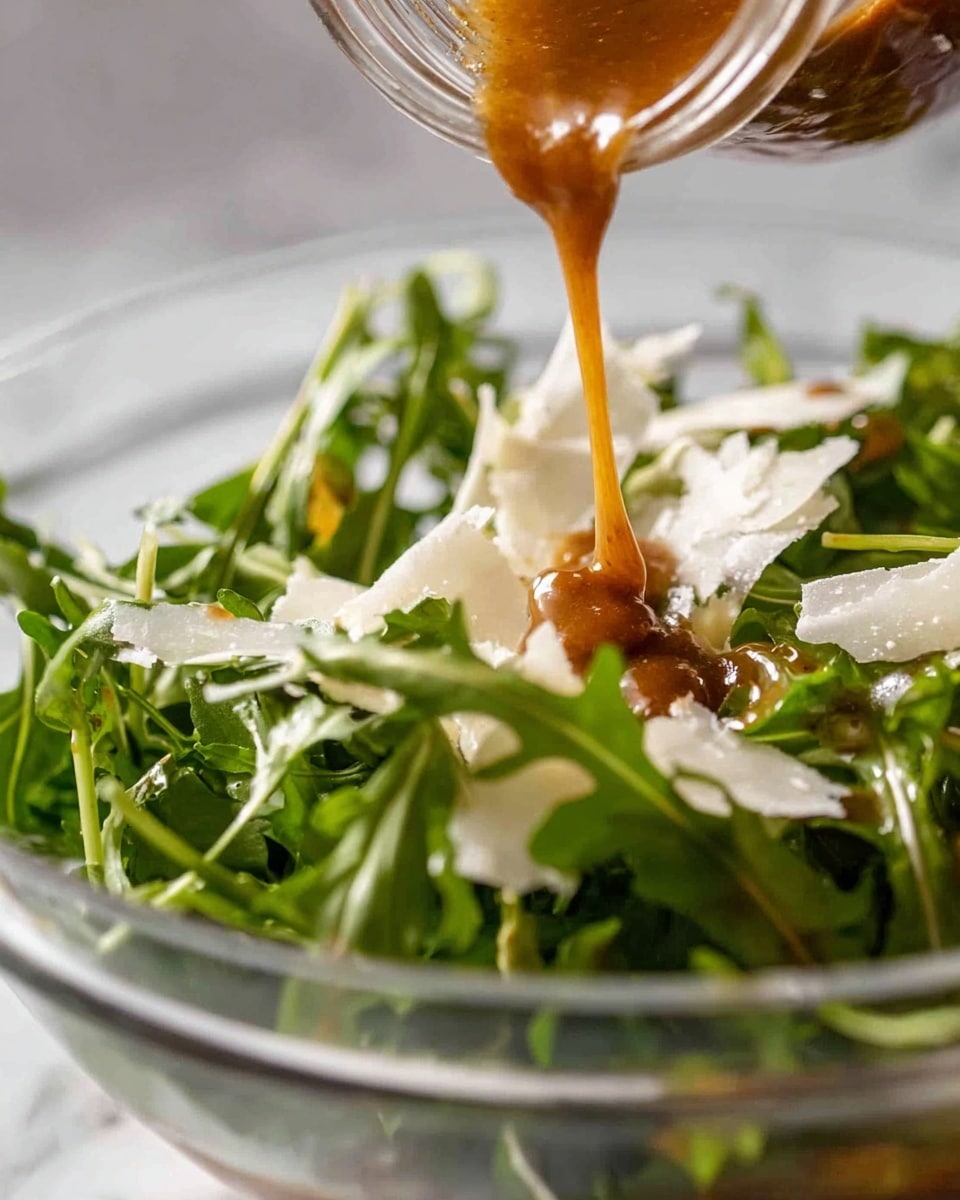 A close-up shot of a fresh green salad in a white bowl on a white marbled surface, showing one layer of dark green arugula leaves with thin, pale white shavings of cheese scattered on top. A thick, glossy brown dressing is being poured from a jar directly onto the salad, dripping down onto the leaves and cheese. The photo captures the texture of the leafy greens and the smooth liquid flow of the dressing. photo taken with an iphone --ar 4:5 --v 7