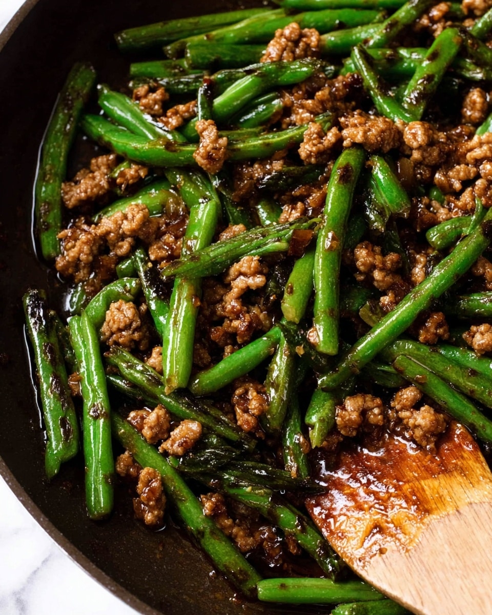 The image shows a close-up of a stir-fry dish with two main layers: bright green beans cut into short pieces, and small chunks of browned ground meat mixed throughout. The green beans have some dark char marks giving a slight contrast to their shiny green skin. The ground meat pieces are coated in a glossy, dark brown sauce that looks thick and sticky, gripping both the beans and meat well. A wooden spoon is scooping some of the stir-fry on the right side, adding a natural texture to the scene. The dish is placed on a black pan with a white marbled texture as the background. Photo taken with an iphone --ar 4:5 --v 7